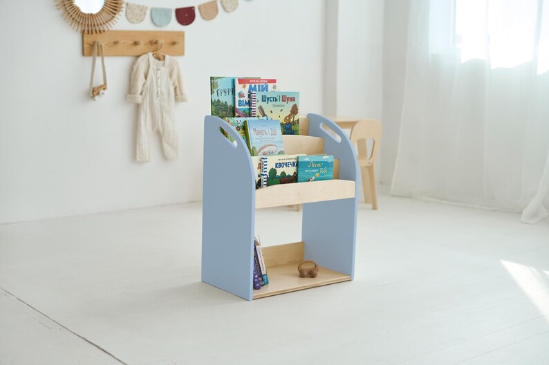 May include: A pale blue wooden bookcase with three shelves, displaying various colourful children's books. The bookcase has a light wood shelf and base. The design includes handles on the sides. The room has a white wall and floor.