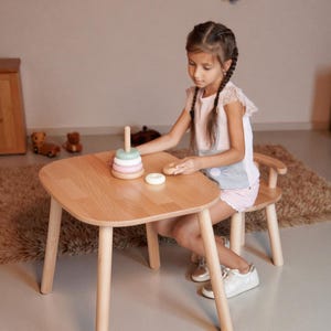 K&ouml;nnte beinhalten: Ein Kindertisch- und Stuhlset aus hellem Holz. Der Tisch hat abgerundete Kanten und vier Beine. Ein Holz-Steckspielzeug mit pastellfarbenen Ringen liegt auf dem Tisch. Ein Kind sitzt am Tisch.