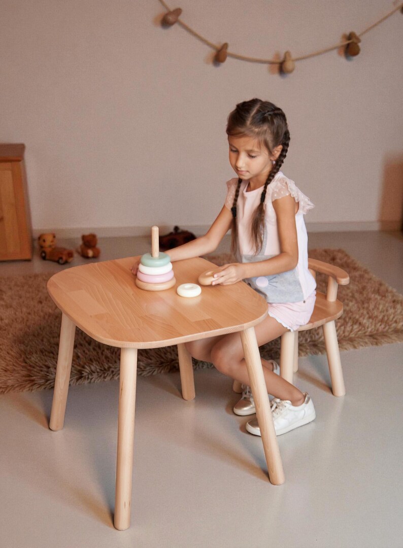 K&ouml;nnte beinhalten: Ein Kindertisch- und Stuhlset aus hellem Holz. Der Tisch hat abgerundete Kanten und vier Beine. Ein Holz-Steckspielzeug mit pastellfarbenen Ringen liegt auf dem Tisch. Ein Kind sitzt am Tisch.
