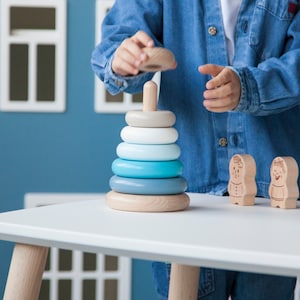 May include: A wooden stacking toy with rings in shades of blue, white, and gray. The toy is on a white table, with two small wooden figures nearby. A person is holding the top ring, ready to place it on the central post.