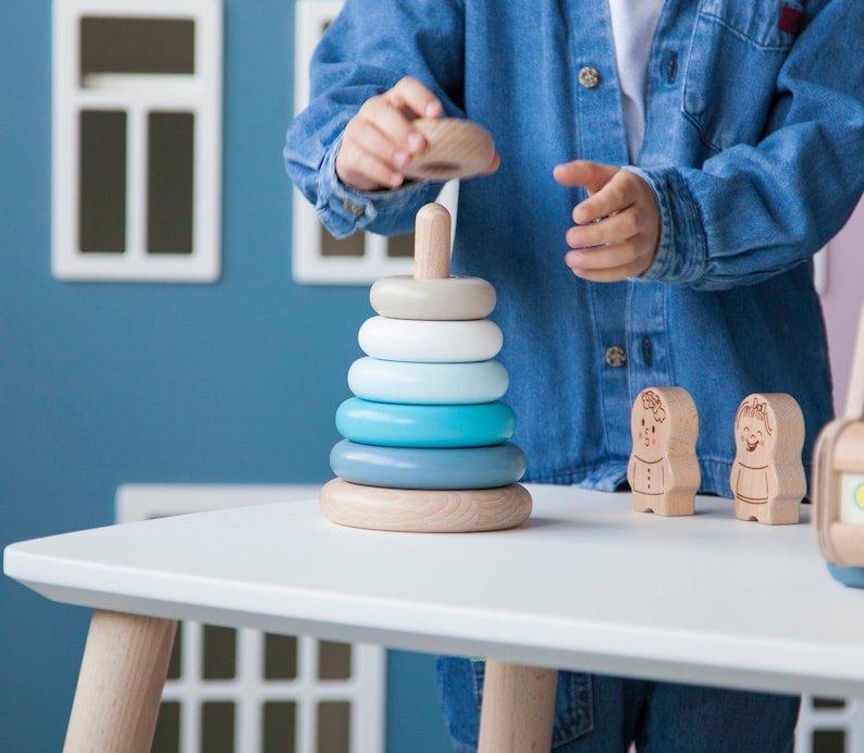 May include: A wooden stacking toy with rings in shades of blue, white, and gray. The toy is on a white table, with two small wooden figures nearby. A person is holding the top ring, ready to place it on the central post.