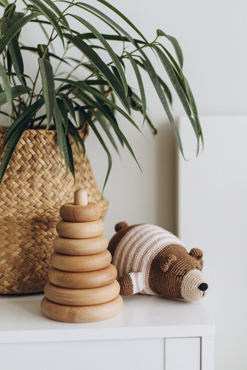 May include: A wooden stacking ring toy with a light brown finish and a crocheted bear toy with a striped shirt. The toys are on a white surface, with a woven basket and a green plant in the background.