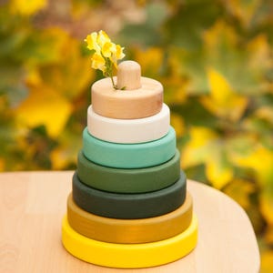 Puede incluir: Un juguete apilable de madera con una parte superior de madera natural y una serie de anillos coloridos en tonos de amarillo, verde y blanco. Una pequeña flor amarilla está colocada en el centro de la pieza superior. El juguete está sobre una superficie de madera clara.