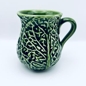 May include: A green ceramic mug shaped like a cabbage. The mug has a handle and a textured surface.