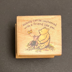 May include: A wooden stamp with a Winnie the Pooh and Piglet illustration. The text reads "Nobody can be uncheered with a friend like you."