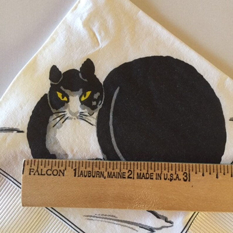 May include: A white fabric napkin with a black and white cat illustration. The cat has yellow eyes and is sitting with its back to the viewer. The napkin has a wooden ruler with the text "FALCON 1 AUBURN, MAINE 2 MADE IN U.S.A. 31" printed on it.