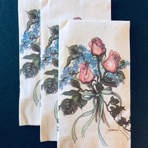 May include: Three white paper napkins with a floral design. The design features three pink roses with green leaves and blue flowers. The napkins have a scalloped edge and the text "CHRISTMAS" is printed on the bottom right corner.