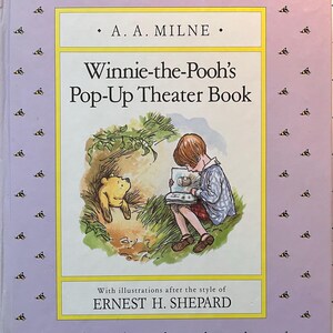 May include: A hardcover book titled Winnie-the-Pooh's Pop-Up Theater Book by A. A. Milne. The cover features a light purple border with small bee icons, a central illustration of Christopher Robin reading to Winnie-the-Pooh, and text credits.