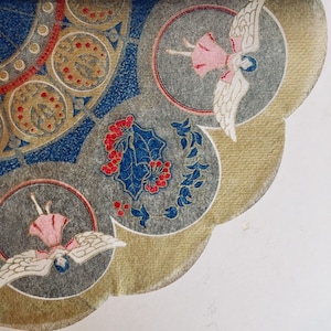 May include: Close-up of a decorative paper design with a repeating pattern. The design features circular motifs with angels, berries, and floral elements in shades of pink, red, blue, gold, and gray. The paper has a scalloped edge.