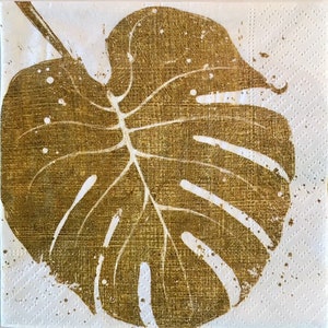 Puede incluir: Una impresión de hoja de oro de una hoja de monstera sobre un fondo blanco.