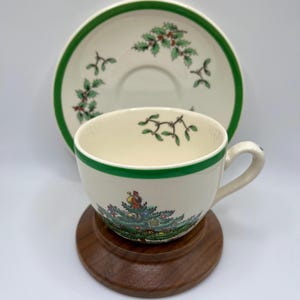 May include: A white teacup and saucer with a green rim and a Christmas tree design. The saucer has a green rim and a holly design. The teacup and saucer are sitting on a wooden stand.