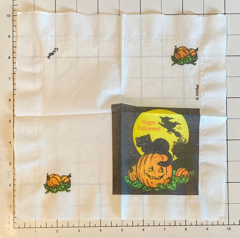 May include: White paper napkin with a Halloween theme. The napkin features a black cat sitting on a jack-o-lantern with a full moon and a witch flying in the background. The text "Happy Halloween!" is printed on the napkin.