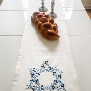 May include: A white linen table runner with a blue embroidered Star of David design. A braided challah bread sits on the runner, and two silver candlesticks with white candles are in the background.