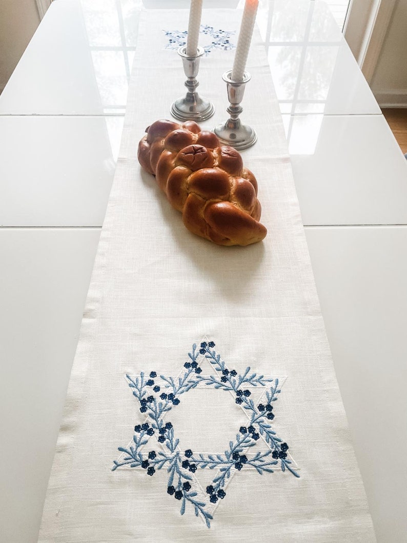 May include: A white linen table runner with a blue embroidered Star of David design. A braided challah bread sits on the runner, and two silver candlesticks with white candles are in the background.