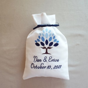 Könnte beinhalten: Weißer Stoff-Geschenkbeutel mit blauem Baumdesign, bestickt mit den Namen "Dan & Erica" und dem Datum "10. Oktober 2021" in Marineblau. Der Beutel ist mit einem marineblauen Band gebunden.