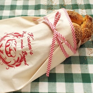 May include: A white cloth bread bag with red and white striped ribbon tied around it. The bag has a red embroidered design with the text "Karen's Home Made Bread". A loaf of bread is inside the bag.