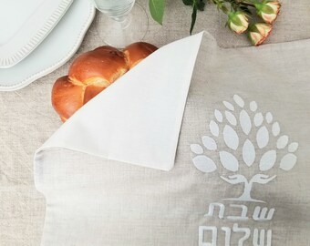 May include: A white linen challah cover with a white embroidered tree design and the Hebrew words "Shalom" and "Shabbat" in white. A challah bread is partially visible under the cover.