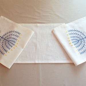 May include: Two white linen table runners with blue and yellow embroidered tree designs. The trees are stylized and have a whimsical look.