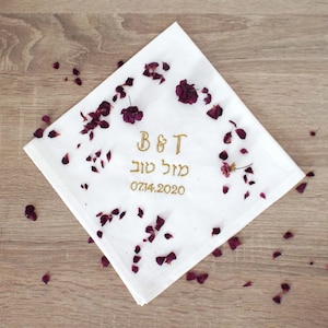 May include: White fabric handkerchief with gold embroidery. The embroidery includes the initials "B&T", the Hebrew words "Mazal Tov", and the date "07.14.2020". The handkerchief is surrounded by dried rose petals.