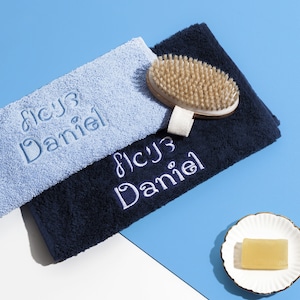 May include: Two personalized towels, one light blue and one navy blue, with the name "Daniel" embroidered on each. A wooden bath brush and a bar of soap are also pictured.