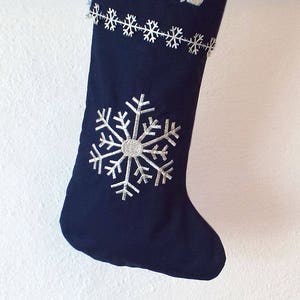 May include: A dark blue stocking with a white snowflake design and the name "Micah" embroidered in white Hebrew letters.