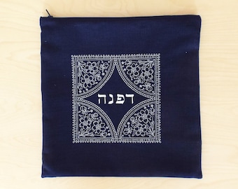 May include: A dark blue fabric pouch with a white embroidered design featuring a floral pattern and the Hebrew word "Shabbat" in the center.