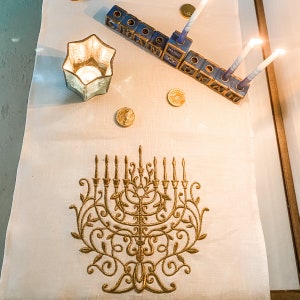 Könnte beinhalten: Eine weiße Tischdecke mit einem goldenen gestickten Menora-Design. Auf dem Tisch befinden sich blaue und goldene Blöcke mit hebräischen Buchstaben, die das Wort 'Chanukka' buchstabieren, sowie brennende Kerzen.