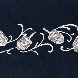 Può includere: Cinque dreidel bianchi ricamati con lettere ebraiche su uno sfondo blu scuro. I dreidel sono disposti in fila con dei riccioli decorativi intorno.