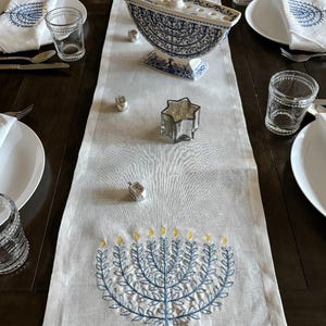 May include: A white linen table runner with blue and yellow embroidered menorahs. The runner is decorated with a blue and white ceramic menorah and silver dreidels.