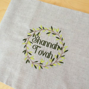 Puede incluir: Un mantel individual de lino beige con una corona de ramas de olivo bordada y el texto "Shannah Tovah" en negro.