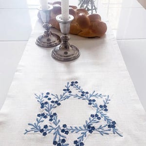 Può includere: Un runner da tavolo in lino bianco con una stella di David ricamata in blu. La stella è circondata da una ghirlanda di foglie e fiori blu. Due candelabri d'argento con candele bianche sono sul runner da tavolo.