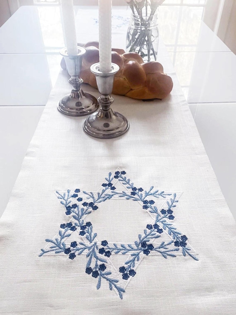 May include: White linen table runner with a blue embroidered Star of David. The star is surrounded by a wreath of blue leaves and flowers. Two silver candle holders with white candles are on the table runner.