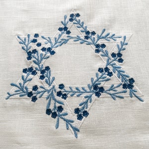 May include: A white fabric with a blue embroidered Star of David. The star is outlined with blue leaves and small blue flowers.