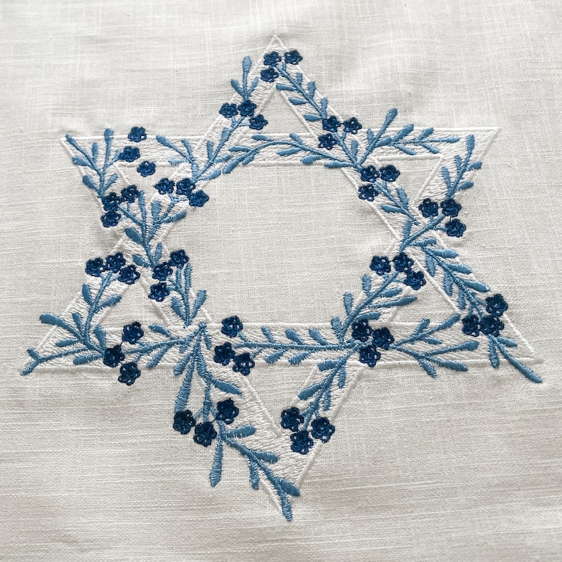 May include: A white fabric with a blue embroidered Star of David. The star is outlined with blue leaves and small blue flowers.