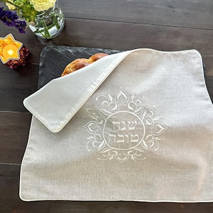 May include: A beige linen challah cover with a white embroidered floral design and the Hebrew text "Shabbat Shalom". The cover has a white border and is partially covering a loaf of bread on a black serving board.