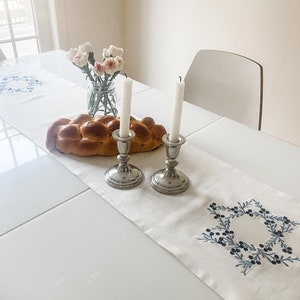 May include: A white table runner with a blue embroidered Star of David design. Two silver candle holders with white candles are on the runner, along with a challah bread and a vase of pink flowers.