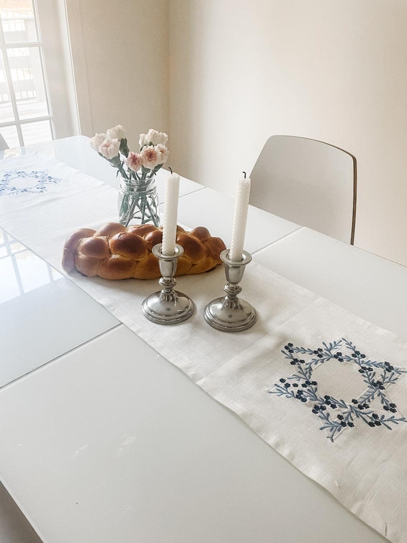 May include: A white table runner with a blue embroidered Star of David design. Two silver candle holders with white candles are on the runner, along with a challah bread and a vase of pink flowers.