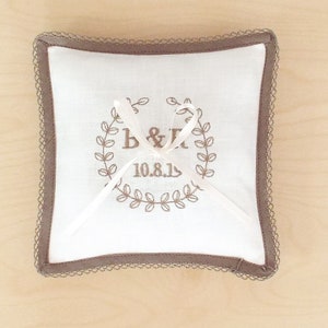May include: Square white ring pillow with brown embroidered details and a decorative border. The pillow features a wreath design with the initials "B & R" and the date "10.8.13". A white ribbon is tied on top.