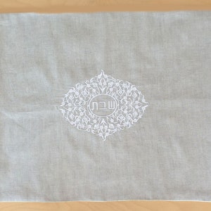 May include: A gray linen fabric challah cover with white embroidery featuring a floral design and the Hebrew word "Shabbat" in the center.