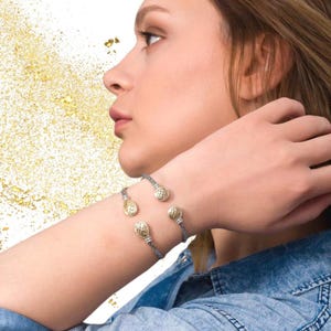 May include: A gray braided bracelet with three gold-toned charms. The charms have a delicate, intricate design.