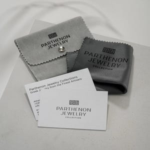 Może przedstawiać: Biała karta z tekstem "Parthenon Jewelry Collection" i tekstem "Greek Jewelry from the Finest Artisans" nadrukowanym na niej. Karta leży na szarej aksamitnej torebce z nadrukiem "Parthenon Jewelry".