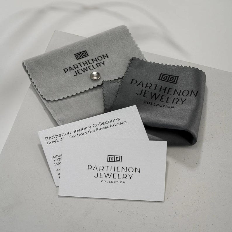 Może przedstawiać: Biała karta z tekstem "Parthenon Jewelry Collection" i tekstem "Greek Jewelry from the Finest Artisans" nadrukowanym na niej. Karta leży na szarej aksamitnej torebce z nadrukiem "Parthenon Jewelry".