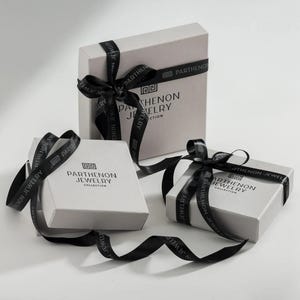 Może przedstawiać: Trzy białe pudełka prezentowe z czarną wstążką i tekstem "Parthenon Jewelry Collection" na każdym pudełku.