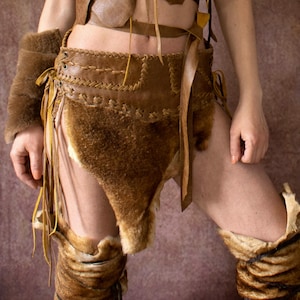 May include: A handmade, brown leather and fur costume. The outfit includes a fur-lined arm cuff, a skirt with leather detailing, and fur-trimmed leg warmers. The costume is designed with a rustic, tribal aesthetic.