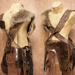 Op de afbeelding: Twee mannequins tonen een handgemaakt, rustiek kostuum. Het kostuum heeft een bontkraag, leren riemen en bontaccenten. Het kleurenpalet omvat bruin- en beigetinten, met de nadruk op natuurlijke materialen en texturen. Het ontwerp suggereert een tribaal of historisch thema.