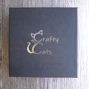 K&ouml;nnte beinhalten: Eine schwarze Box mit einem goldenen Folienlogo einer Katze mit dem Text "Crafty Cats" darauf.
