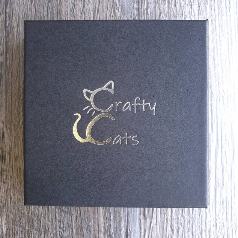 K&ouml;nnte beinhalten: Eine schwarze Box mit einem goldenen Folienlogo einer Katze mit dem Text "Crafty Cats" darauf.
