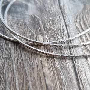 K&ouml;nnte beinhalten: Drei silberne Armreifen mit einer strukturierten, geh&auml;mmerten Oberfl&auml;che. Die Armreifen liegen &uuml;bereinander auf einer Holzoberfl&auml;che.