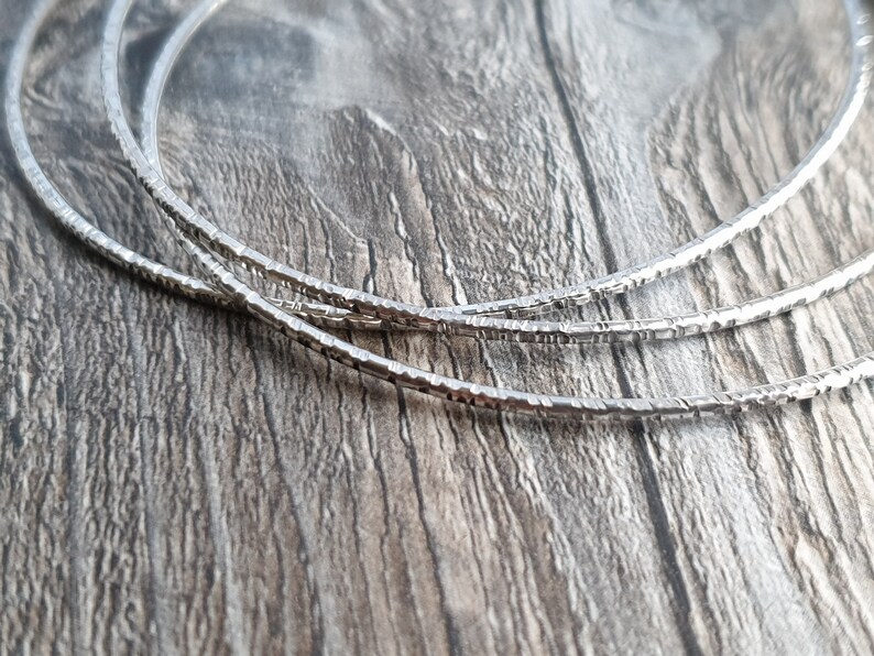 K&ouml;nnte beinhalten: Drei silberne Armreifen mit einer strukturierten, geh&auml;mmerten Oberfl&auml;che. Die Armreifen liegen &uuml;bereinander auf einer Holzoberfl&auml;che.