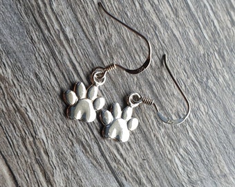 May include: Silver-coloured drop earrings with a paw print design. The earrings have a hook fastening and are shown on a wooden surface. The paw print charms are detailed and three-dimensional.